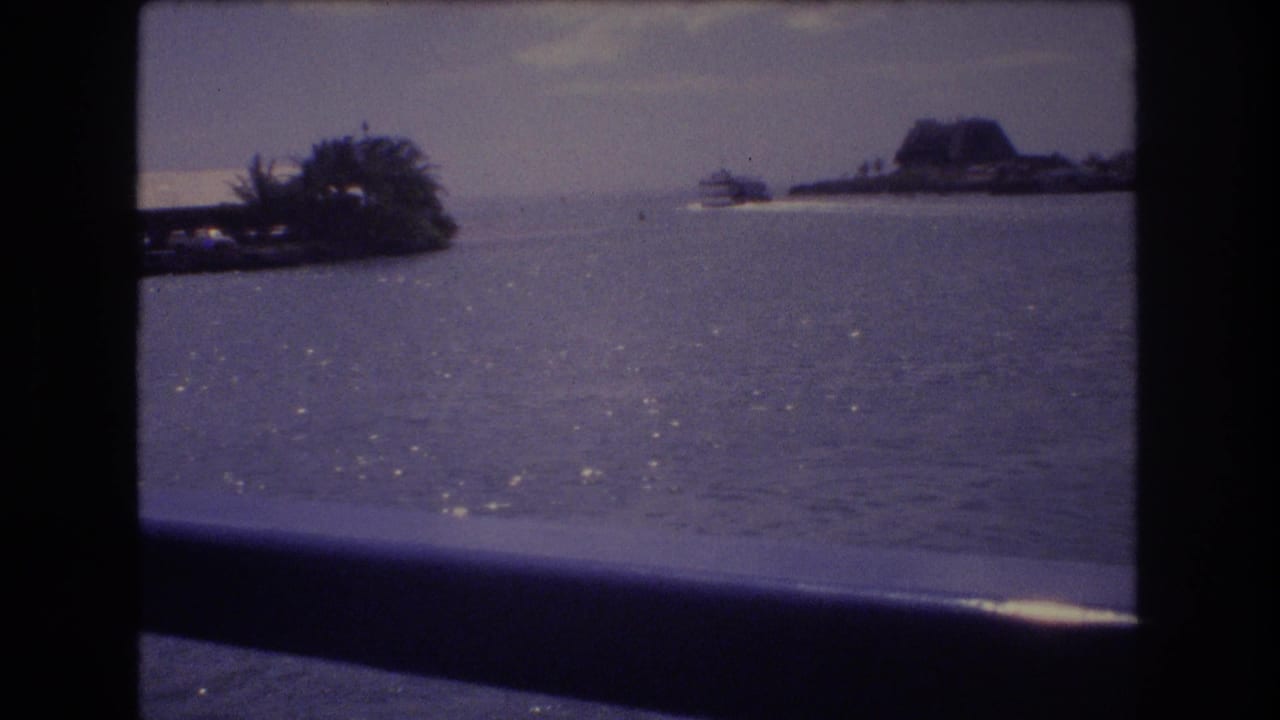 1977 Honolulu home-movie frame looking from a boat across shimmering harbor water toward a low shoreline and distant landform under a muted sky.