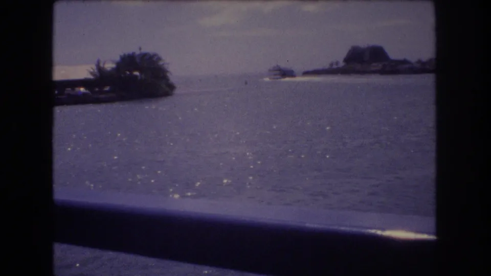 1977 Honolulu home-movie frame looking from a boat across shimmering harbor water toward a low shoreline and distant landform under a muted sky.