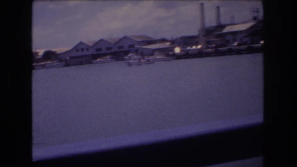 1977 Honolulu home-movie view from a moving boat toward low waterfront buildings, harbor water, and dark railing across the lower frame.