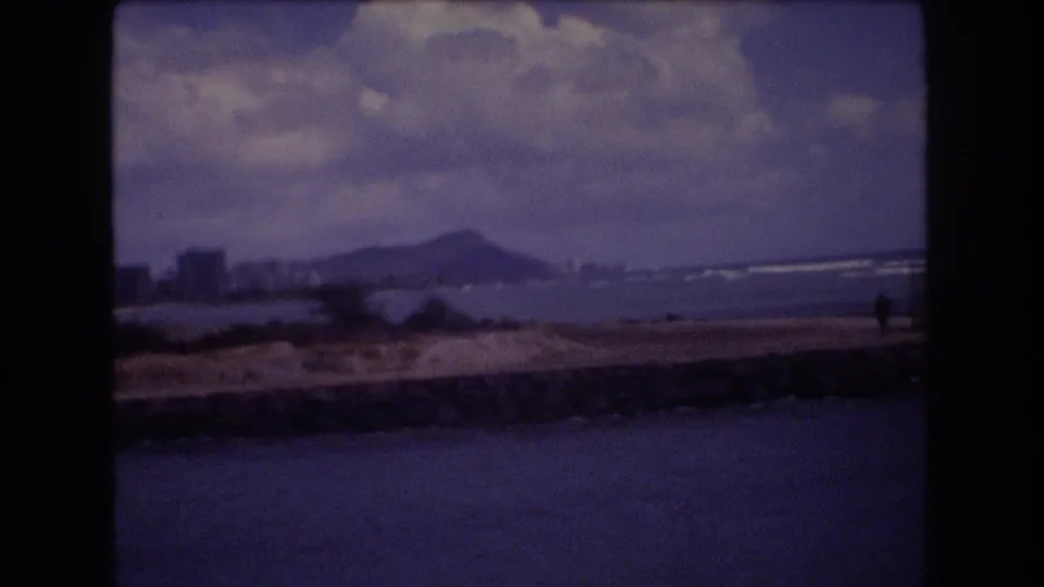1977 Honolulu coastal view with water in the foreground, distant shoreline buildings, mountain profile, cloudy sky, and a small human figure near the right edge.