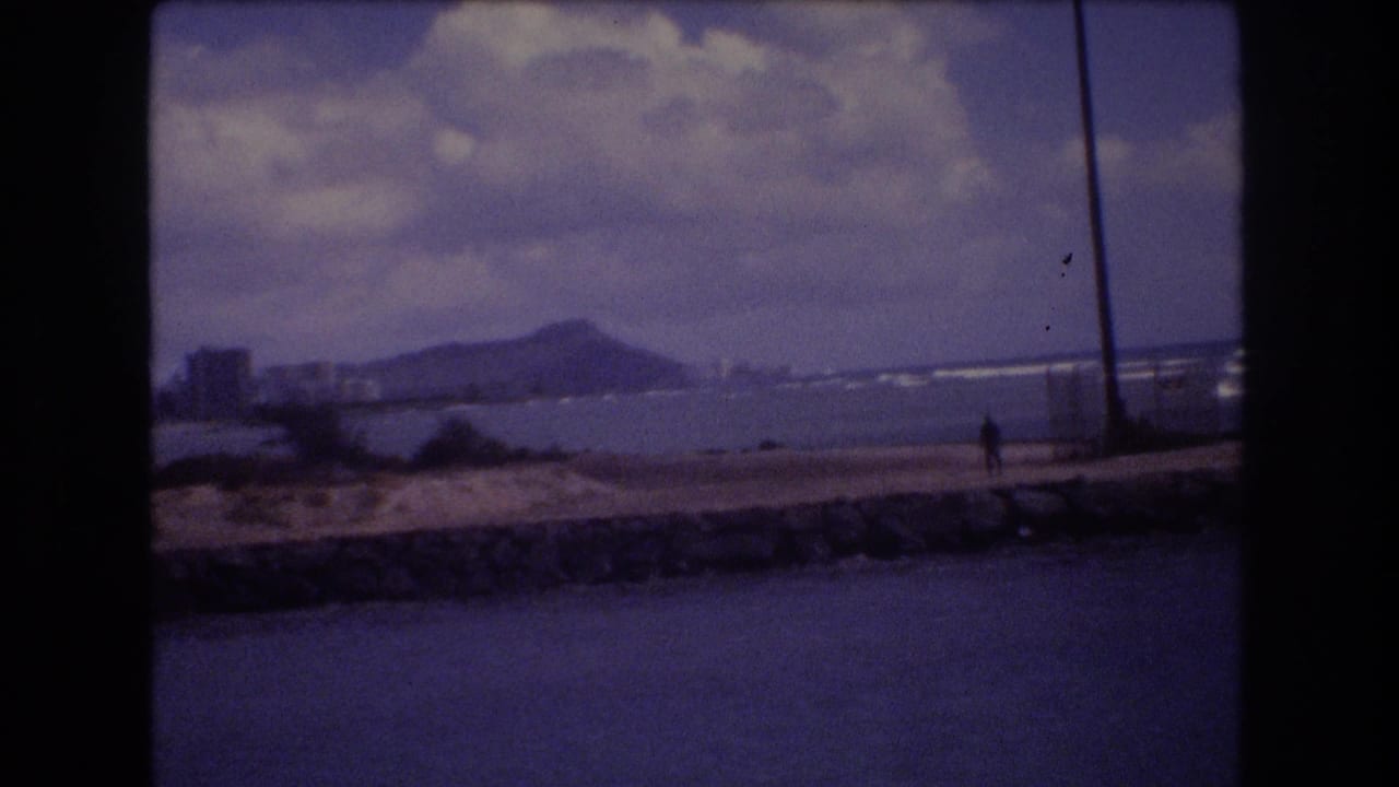 1977 Honolulu home-movie frame showing a lone person on a stone seawall, ocean water in the foreground, and Diamond Head beyond the shoreline.