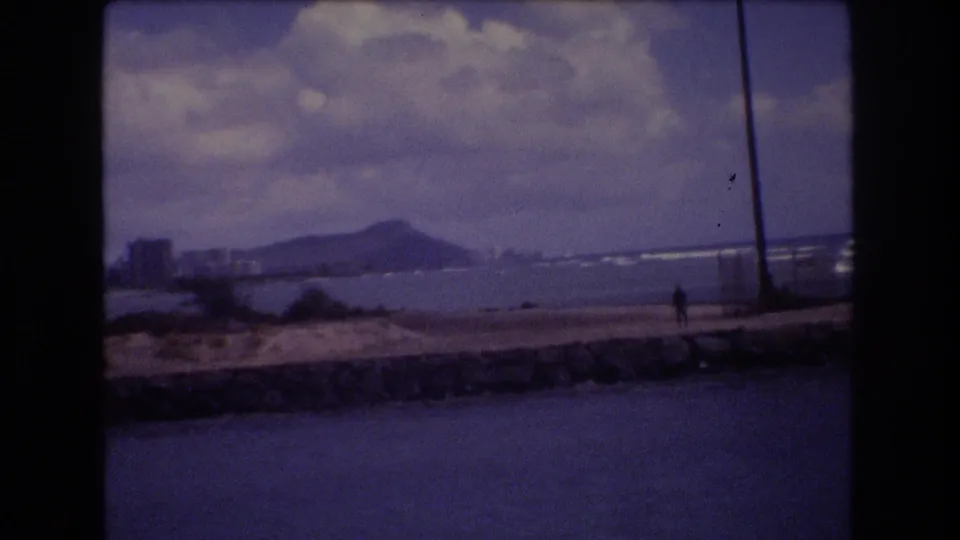 1977 Honolulu home-movie frame showing a lone person on a stone seawall, ocean water in the foreground, and Diamond Head beyond the shoreline.
