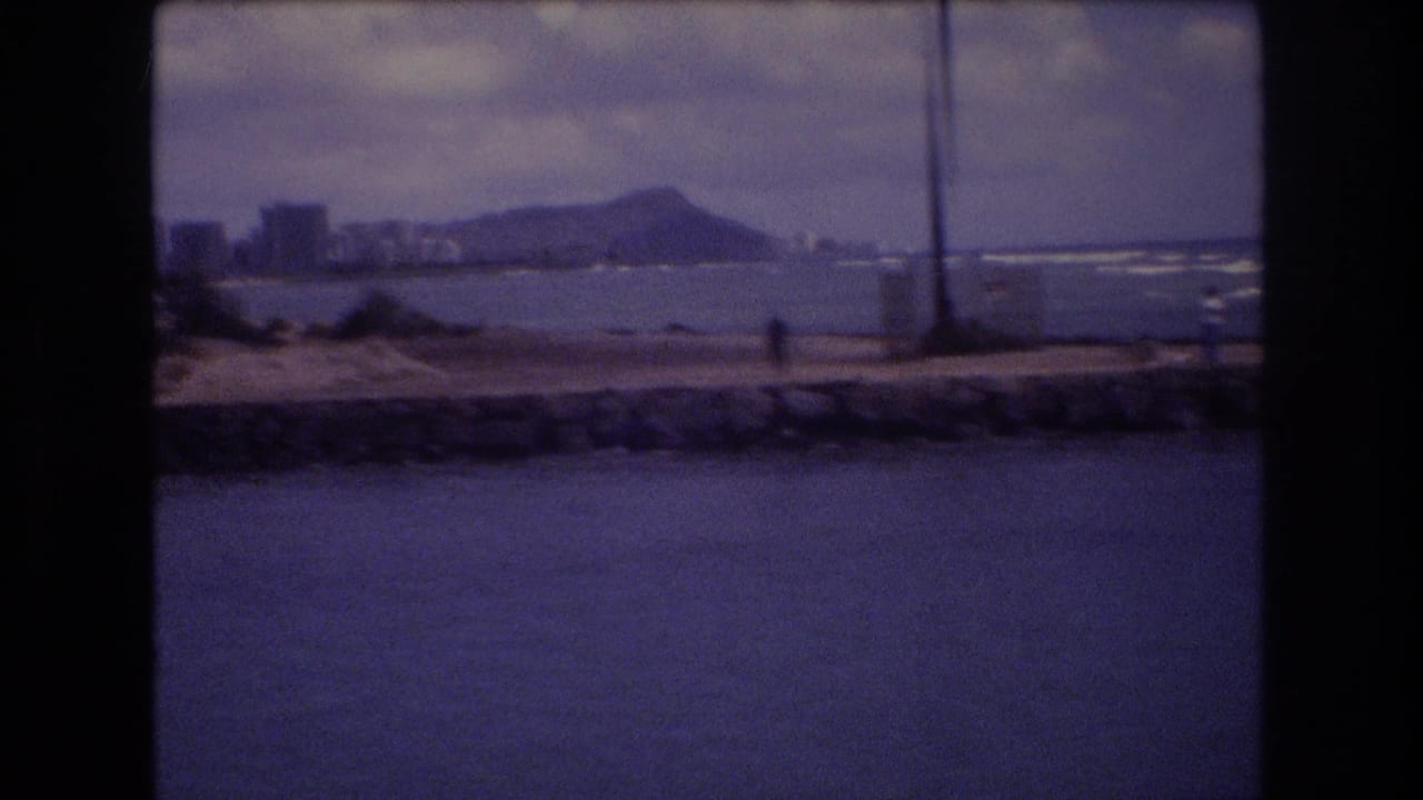 Distant shoreline view with Diamond Head, buildings, seawall, ocean, and a small walking figure.