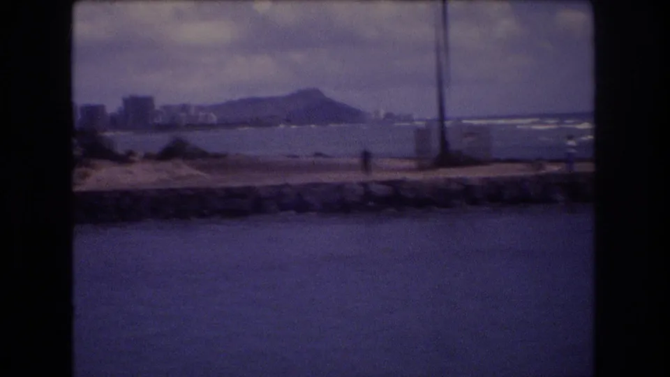 1977 Honolulu coastal frame with Diamond Head behind shoreline buildings, waves breaking near the beach, and a distant person along the seawall.