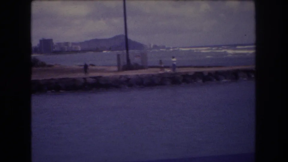 1977 Honolulu frame showing a person near the seawall, Diamond Head and shoreline buildings in the background, and a vertical pole crossing the view.