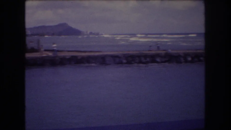1977 Honolulu home-movie frame with open water in the foreground, a stone seawall, distant shoreline figures, and Diamond Head under a cloudy sky.