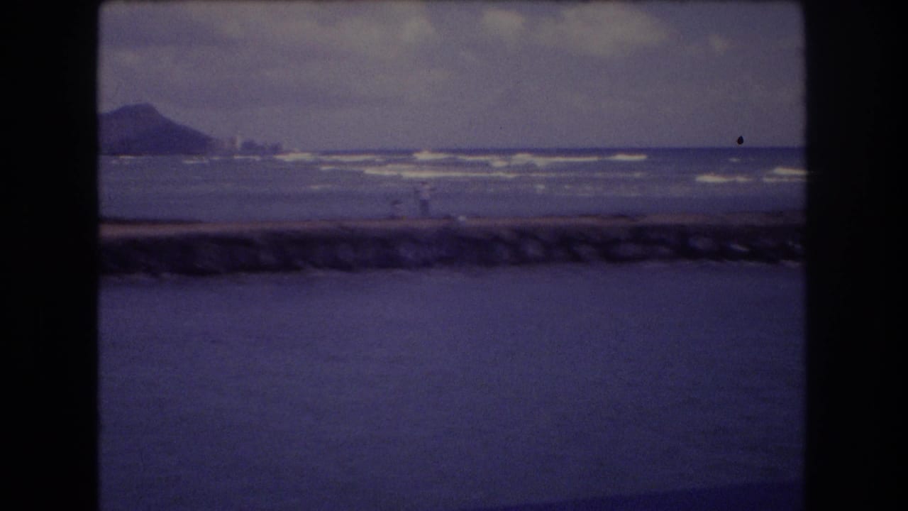 1977 Honolulu 8mm frame showing a rocky breakwater, surf lines, distant hill, cloudy sky, and possible tiny shoreline figures.