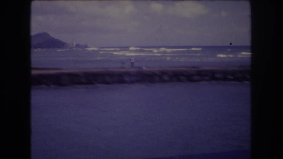 1977 Honolulu 8mm frame showing a rocky breakwater, surf lines, distant hill, cloudy sky, and possible tiny shoreline figures.