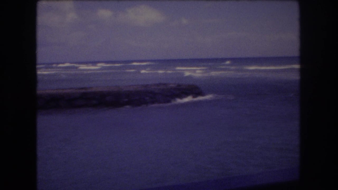 Ocean waves breaking beyond a dark rocky jetty beneath a cloudy sky.