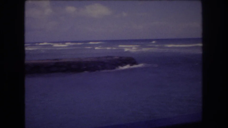 1977 Honolulu 8mm frame with a dark rocky jetty extending into blue ocean water as surf breaks in the distance under cloud cover.
