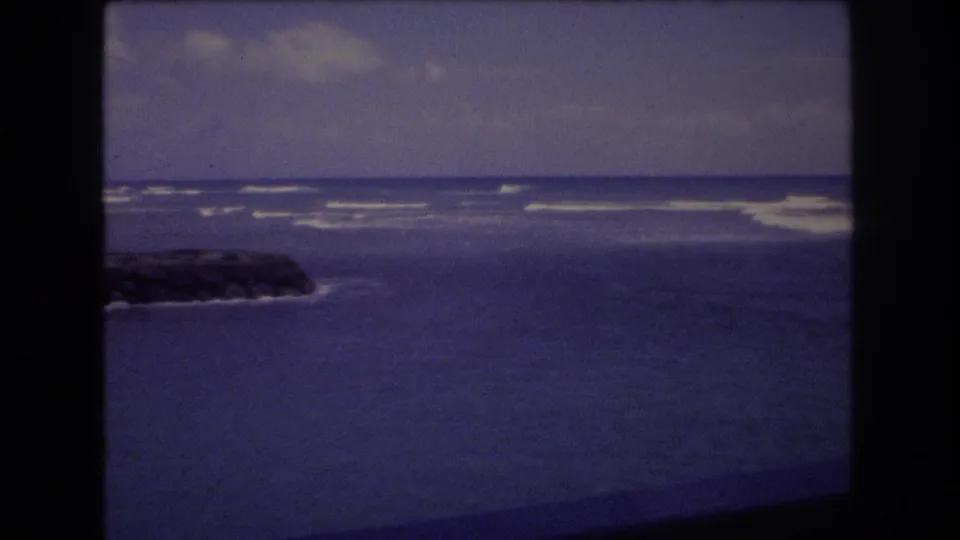 1977 Honolulu 8mm frame showing a low rocky breakwater at the edge of calm water, with white surf bands and a cloudy horizon beyond.