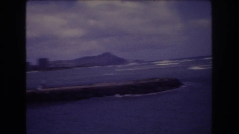 1977 Honolulu 8mm frame with a rocky jetty in the foreground, ocean waves, a distant hill profile, and cloudy sky rendered in soft purple-blue film color.