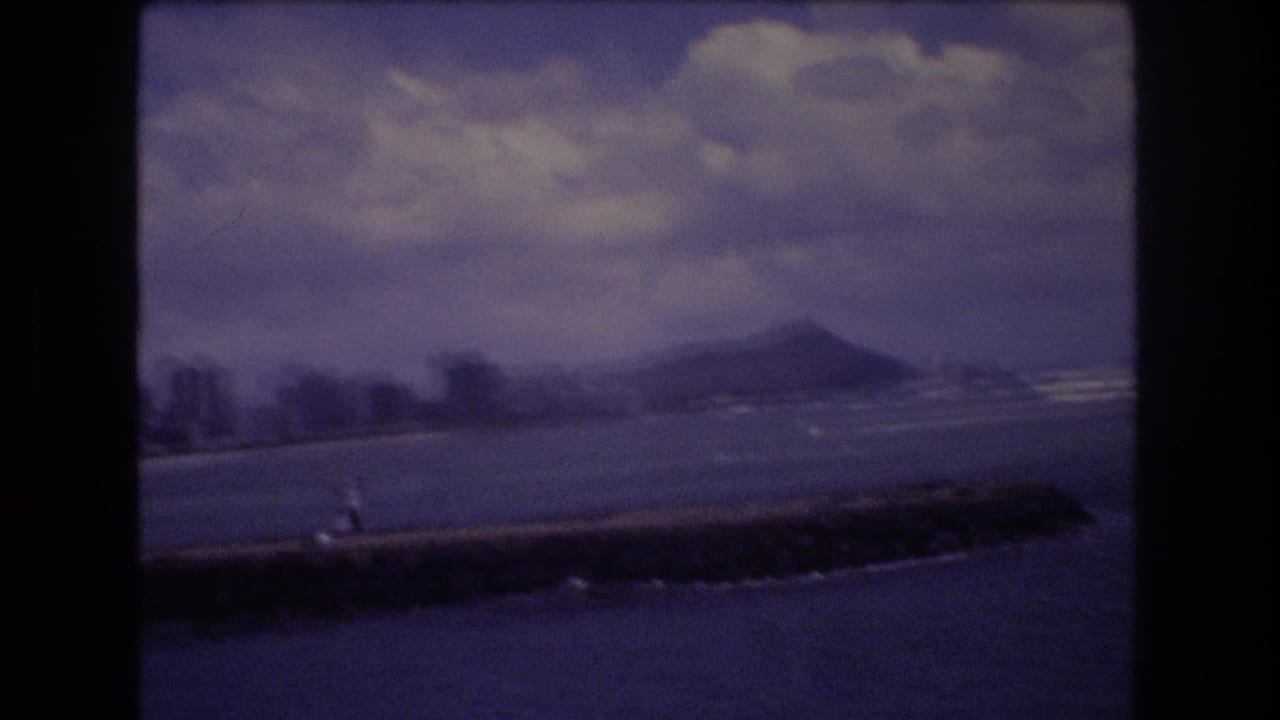 1977 8mm view of Diamond Head beyond Honolulu waters, with a low seawall in the foreground and clouds over the coastline.