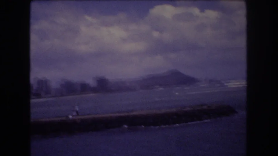 1977 8mm view of Diamond Head beyond Honolulu waters, with a low seawall in the foreground and clouds over the coastline.