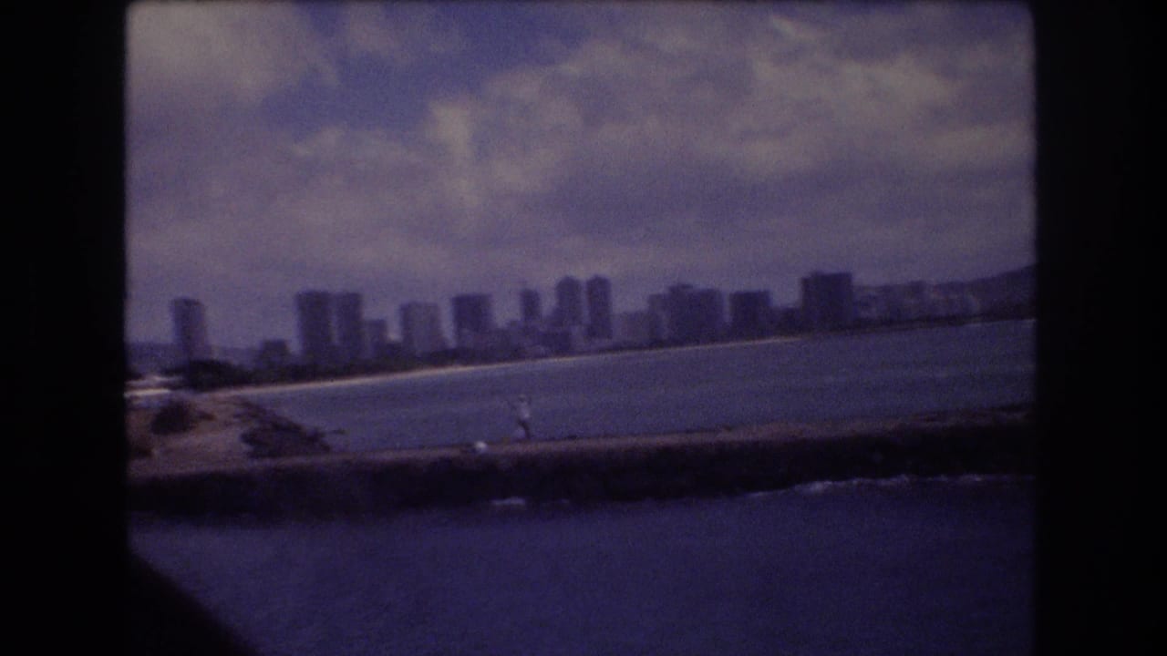 1977 Honolulu waterfront skyline seen across blue water, with high-rise buildings along the shore and a seawall crossing the foreground.