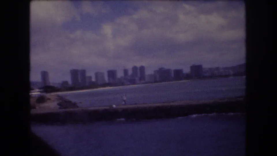 1977 Honolulu waterfront skyline seen across blue water, with high-rise buildings along the shore and a seawall crossing the foreground.