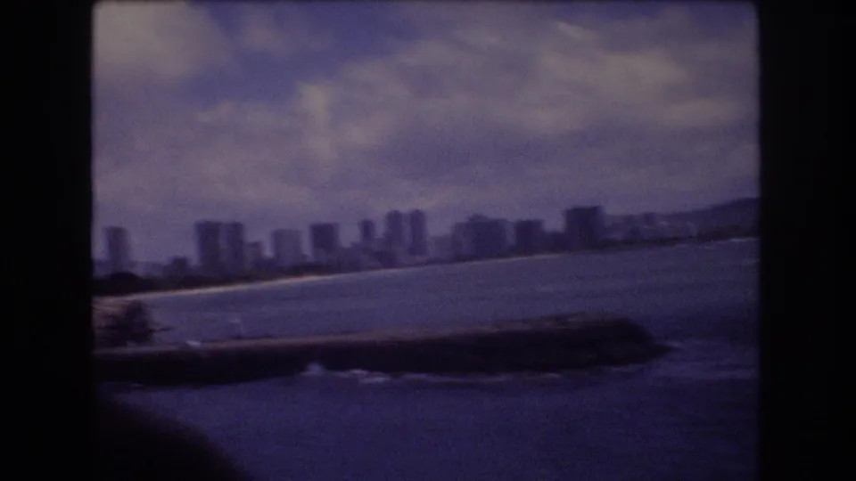 1977 8mm pan frame showing Honolulu buildings along the coast, water in the foreground, and a low seawall or promenade at the edge.