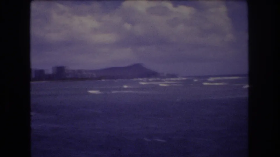 1977 Honolulu view with Diamond Head in the distance, open water in the foreground, and a cloudy sky over the shoreline.