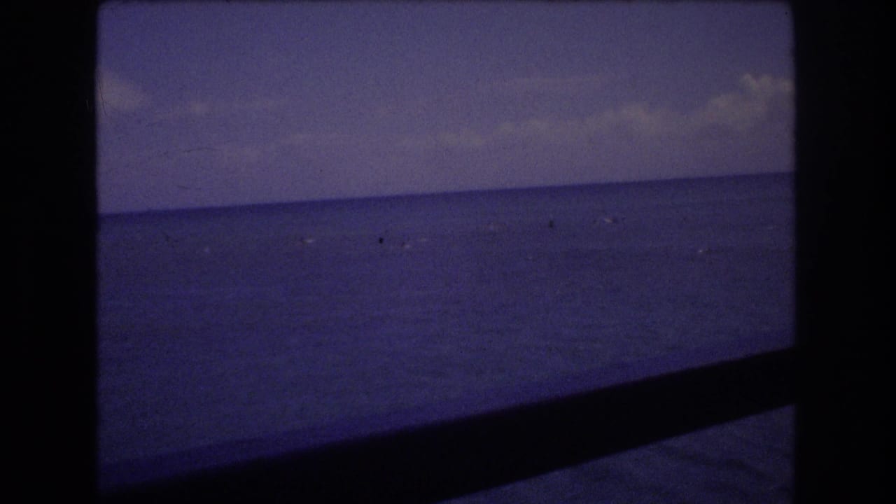 1977 Honolulu 8mm home-movie frame showing a blue ocean horizon under a cloudy sky, with small distant watercraft or specks and dark film-frame borders.