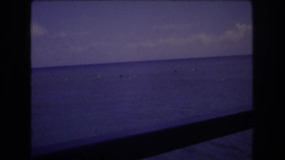 1977 Honolulu 8mm home-movie frame showing a blue ocean horizon under a cloudy sky, with small distant watercraft or specks and dark film-frame borders.