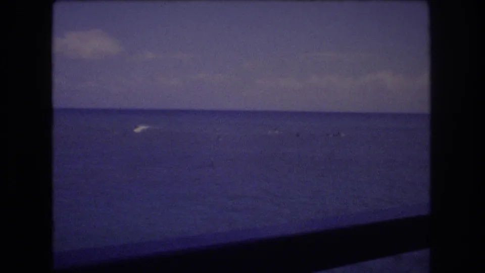 1977 Honolulu 8mm frame of open ocean with a small white wake or breaking water near the horizon beneath a cloudy sky.