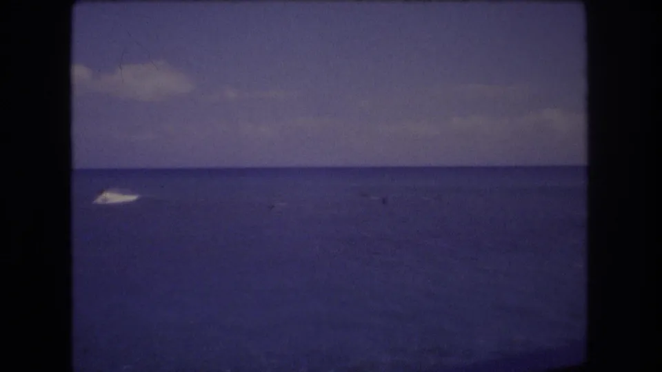 1977 Honolulu home-movie frame showing a blue ocean horizon with a patch of white foam or boat wake entering from the left under soft clouds.
