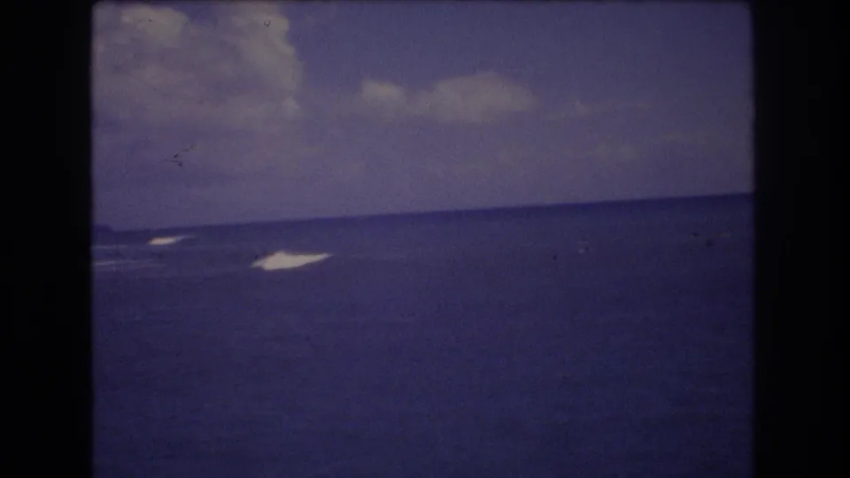 1977 Honolulu 8mm frame showing open ocean with multiple white wakes or foam patches, distant dark specks on the water, and a cloudy sky.