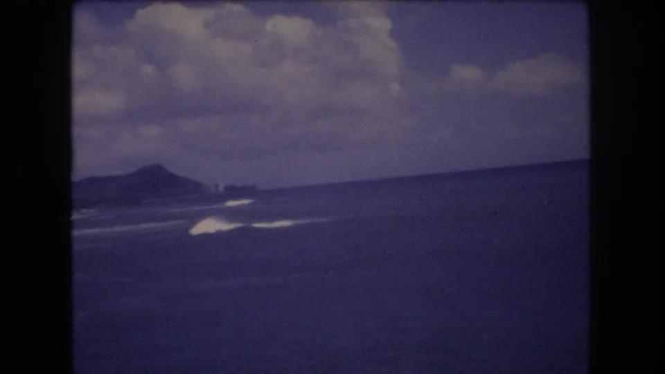 1977 Honolulu coastal view with a distant landmass on the horizon, white surf lines, open blue water, and cloud cover in a soft 8mm frame.