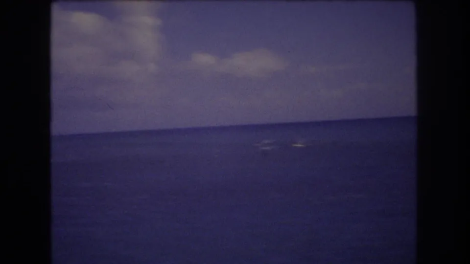 Vintage 8mm Honolulu seascape frame showing a broad ocean surface, faint white surf near the horizon, and a partly cloudy sky.