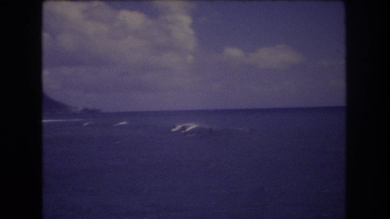 1977 Honolulu 8mm frame showing distant surfers on blue ocean waves with a low coastal ridge on the horizon.