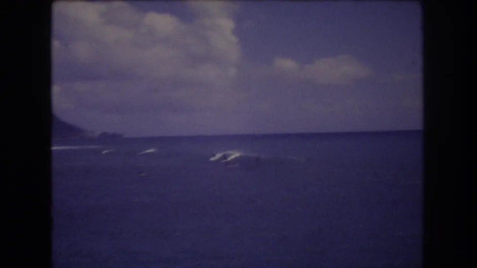 1977 Honolulu 8mm frame showing distant surfers on blue ocean waves with a low coastal ridge on the horizon.