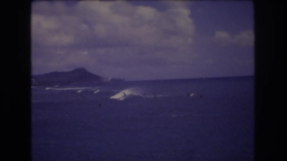 1977 Honolulu 8mm frame with surfers near breaking waves and a distant ridge that appears consistent with Diamond Head.