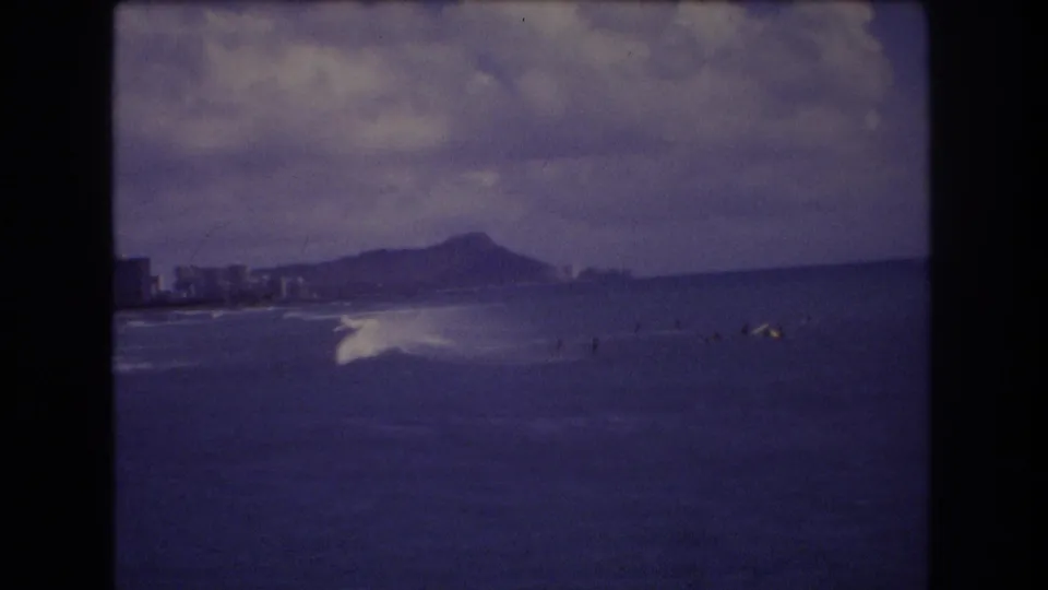 1977 Honolulu 8mm frame showing a breaking wave, small surfers offshore, shoreline buildings, and a Diamond Head-like silhouette.