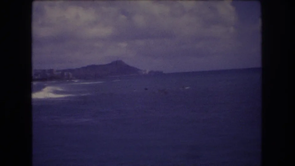1977 Honolulu 8mm frame looking across the ocean toward distant surfers, shoreline development, and a Diamond Head-like ridge.