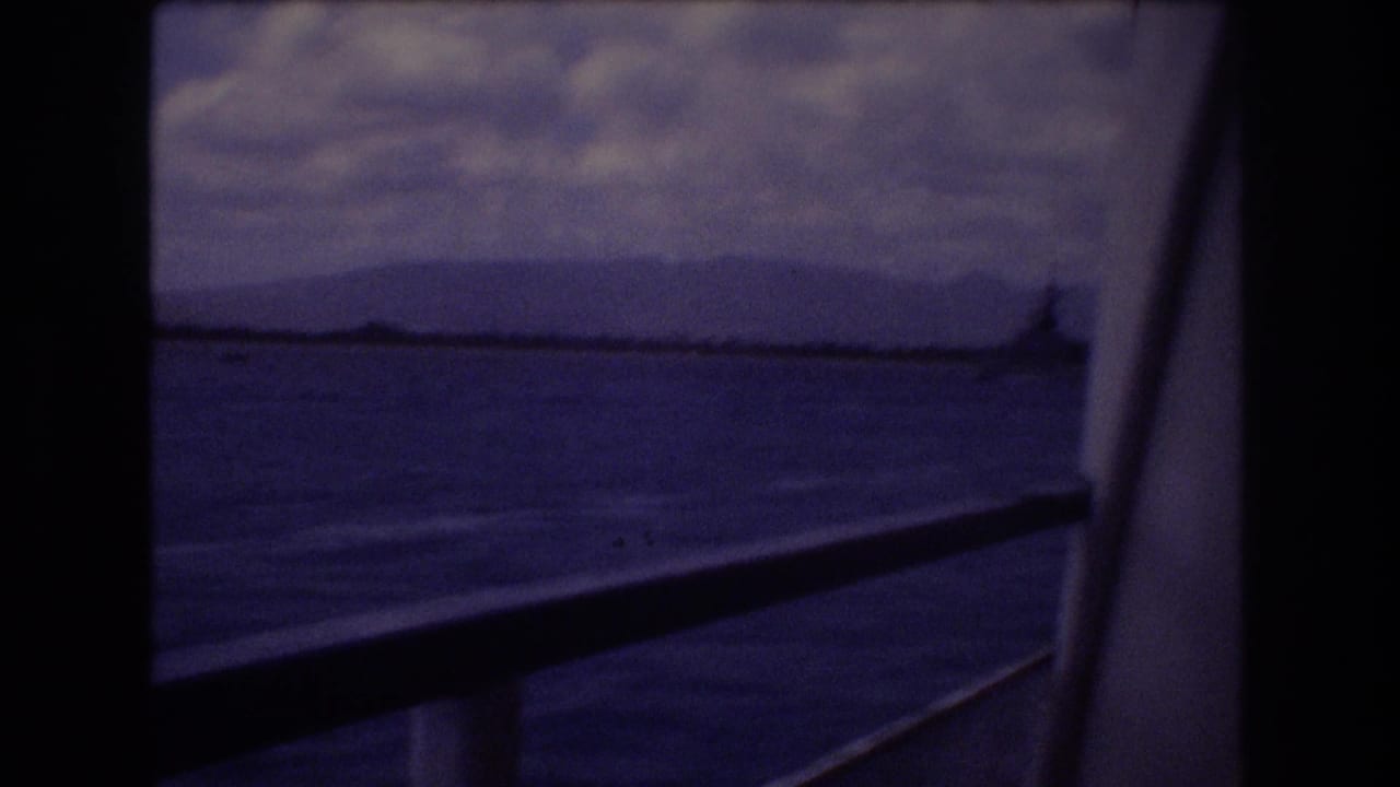 1977 Honolulu home-movie frame showing blue ocean, distant mountain shoreline, cloudy sky, and boat window bars with a railing crossing the foreground.
