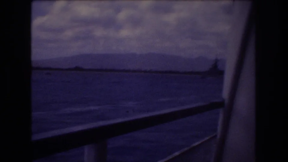 1977 Honolulu home-movie frame showing blue ocean, distant mountain shoreline, cloudy sky, and boat window bars with a railing crossing the foreground.