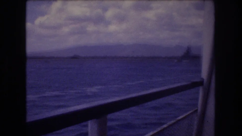 1977 Honolulu boat view with window framing, rippled ocean, distant mountains, cloudy sky, and a large naval-looking ship near the horizon.