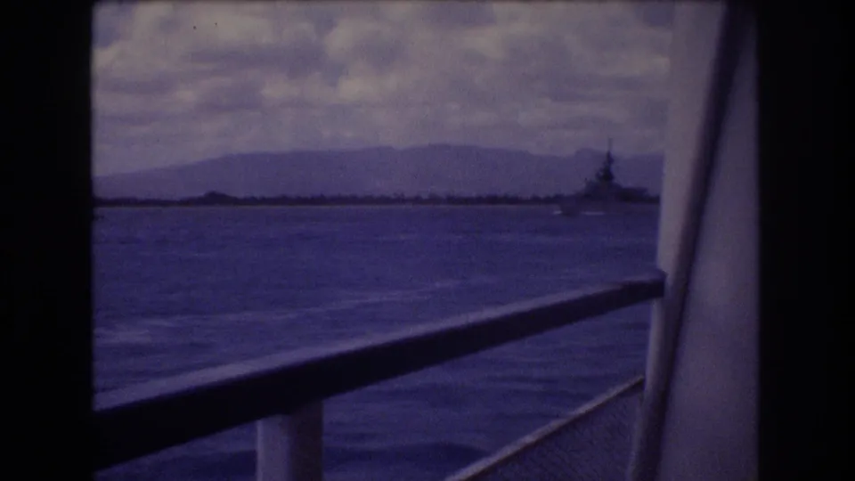 1977 Honolulu harbor-area frame showing a distant naval-looking ship, blue water, mountain backdrop, cloudy sky, and dark boat-window framing.