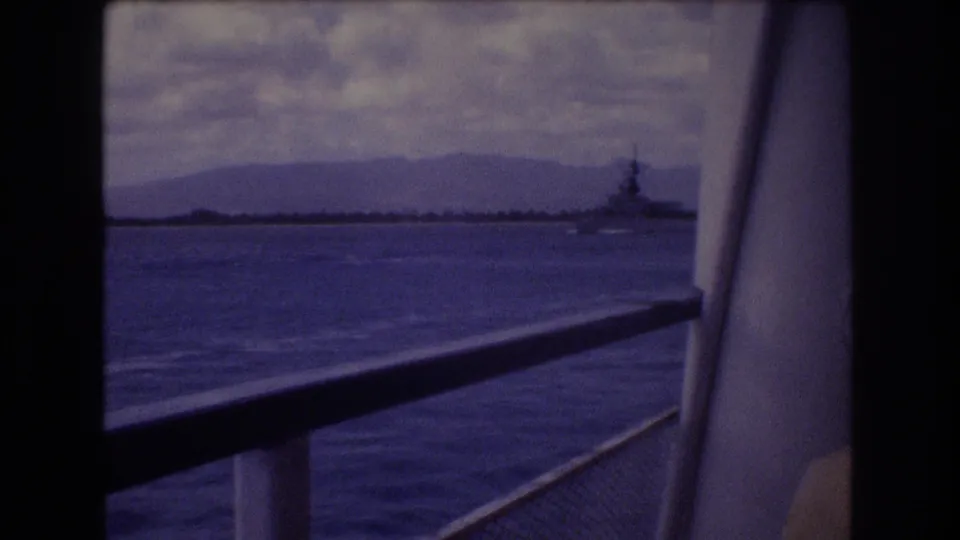 1977 Honolulu home-movie still with dark window bars, boat railings, rippled water, distant mountains, cloudy sky, and a naval-looking ship near shore.