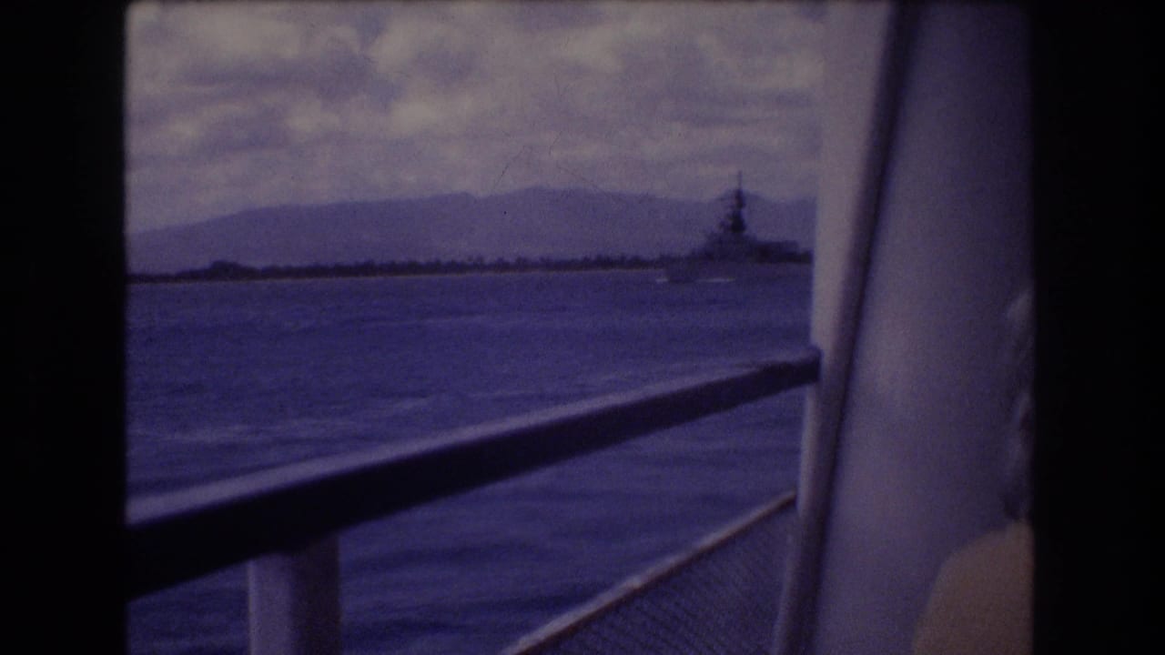 1977 Honolulu-area 8mm frame showing a view from a ship deck across blue water toward a distant naval vessel and low mountains under cloud cover.