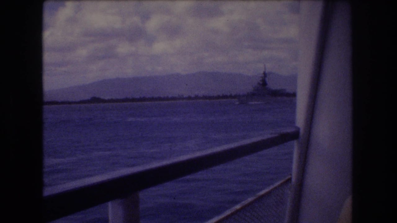 Water seen past a ship railing with mountains and a distant naval vessel on the horizon.