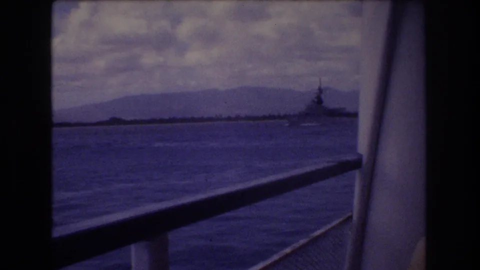 1977 Honolulu-area home-movie frame from a moving ship, with railing in the foreground, water below, and a distant military-looking vessel against mountains.
