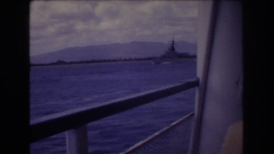 1977 Honolulu-area 8mm frame with purple-blue cast, showing a deck-side view of water, low mountains, and a distant naval vessel near the horizon.
