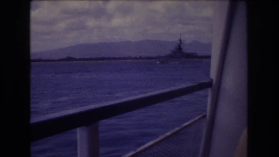 1977 Honolulu-area frame from a ship deck, showing foreground railing, open water, clouded mountains, and a distant military-looking vessel.