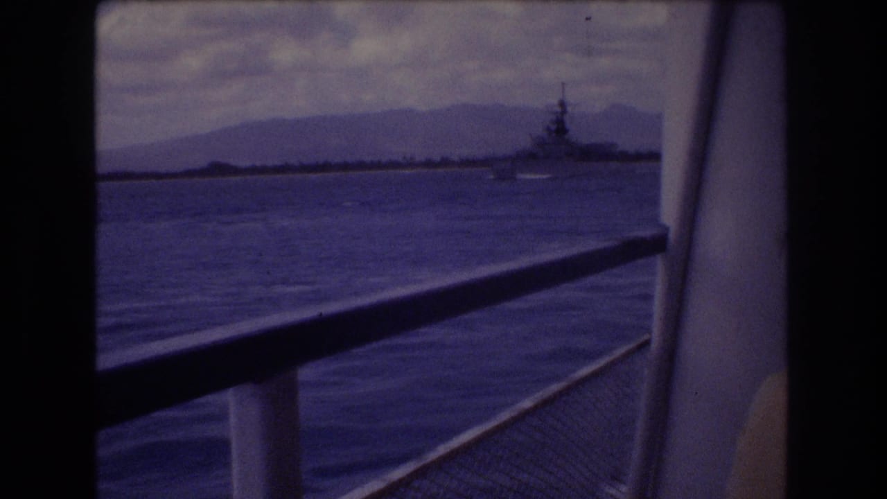 1977 Honolulu 8mm frame showing a distant large vessel on blue water beyond a boat railing, with hazy mountains and cloudy sky in the background.