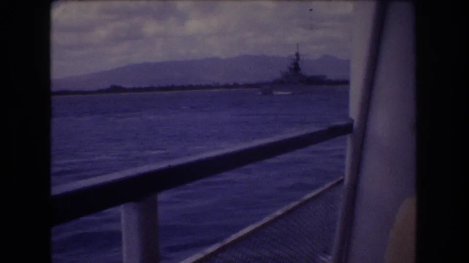 1977 Honolulu 8mm frame showing a distant large vessel on blue water beyond a boat railing, with hazy mountains and cloudy sky in the background.