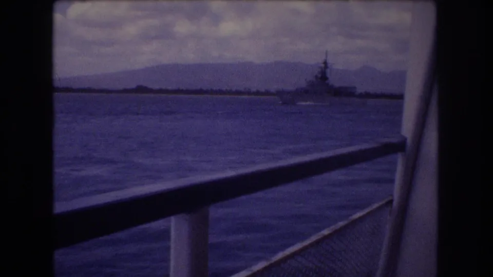1977 Honolulu home-movie frame with a large distant vessel near the horizon, seen from a moving boat through railings and dark frame divisions.