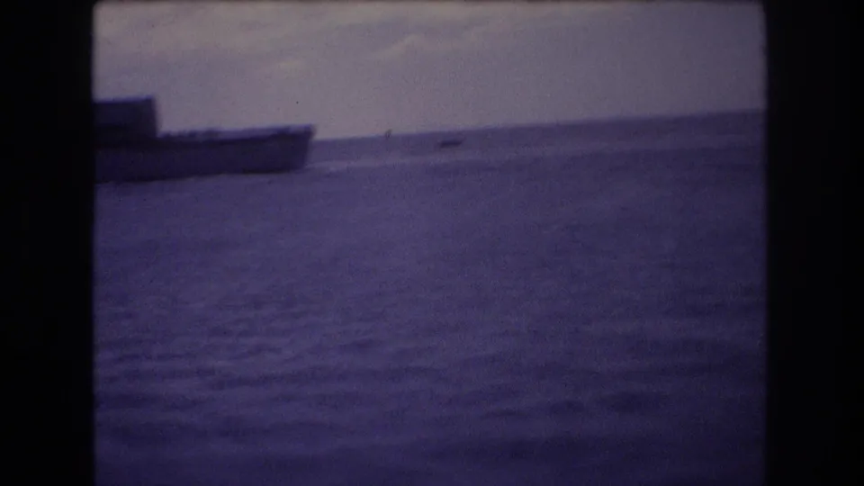 1977 Honolulu 8mm frame showing open water from a boat, with a low vessel at the left edge and faint horizon details under a pale cloudy sky.