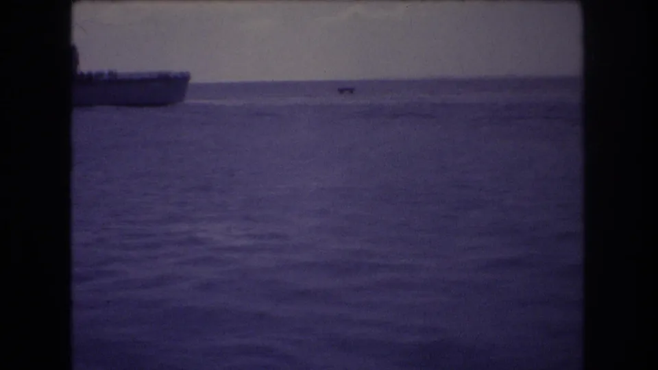 1977 Honolulu home-movie frame looking across calm water, with a low vessel at left, a small distant craft, and a hazy shoreline or mountain profile.
