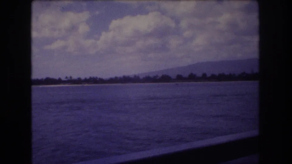 1977 Honolulu coastal view from a moving vantage point, with water in the foreground, distant shoreline and mountains, clouds above, and a dark rail or window edge below.